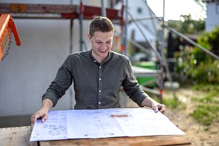Mann steht vor einem Haus mit Baugerüst und schaut auf einen Bauplan
